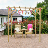 Wooden pergola with chairs and a small table under colorful flags in a garden setting.