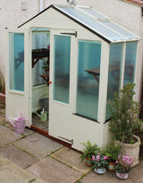 A cream-colored garden shed with frosted glass doors and side windows, set on a stone patio with potted plants and a pink watering can nearby.