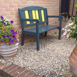A dark teal outdoor bench with bright yellow vertical slats on the backrest, placed on a gravel path against a brick wall with potted flowers nearby.