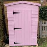 A small pink wooden garden shed with a gabled roof and black metal door hinges, photographed outdoors against a wooden fence and garden background.